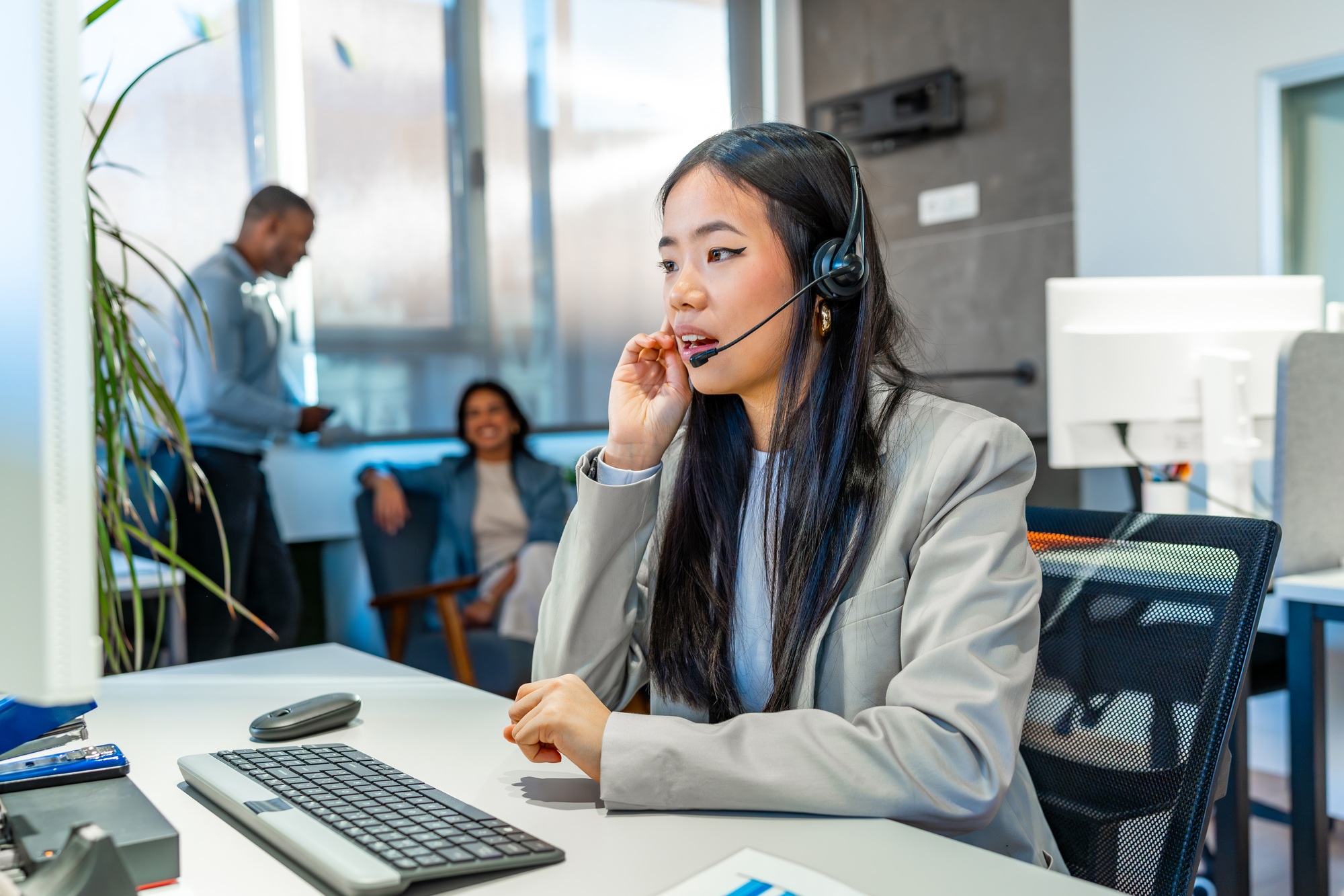 A professional engaged in admin staffing in Los Angeles, wearing a headset and blazer, works in a modern office with colleagues collaborating in the background near large windows and plants.