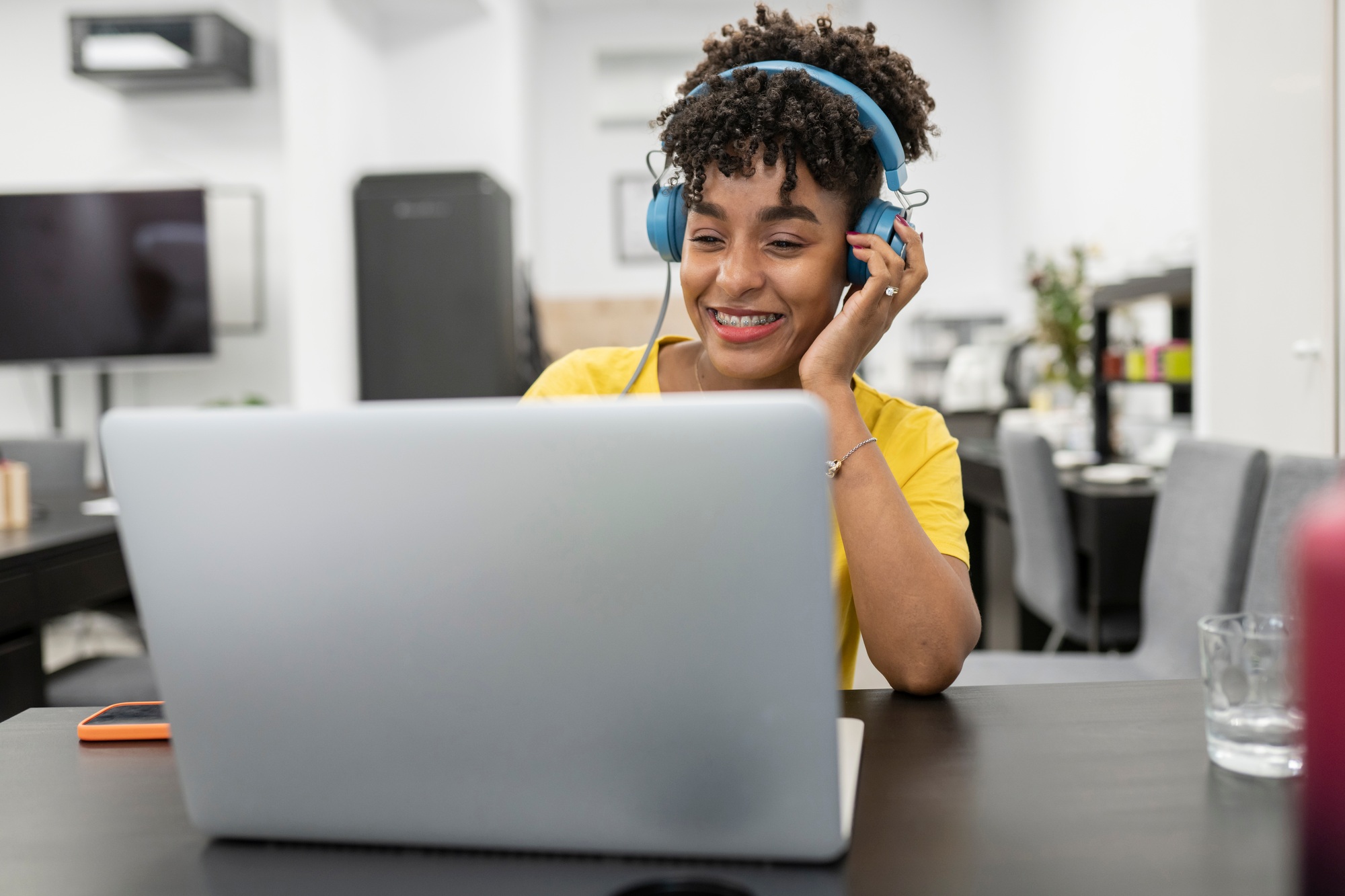 A virtual admin assistant in Los Angeles, wearing a yellow shirt and blue headphones, smiles while working on a laptop in a modern office with a TV and meeting area in the background.