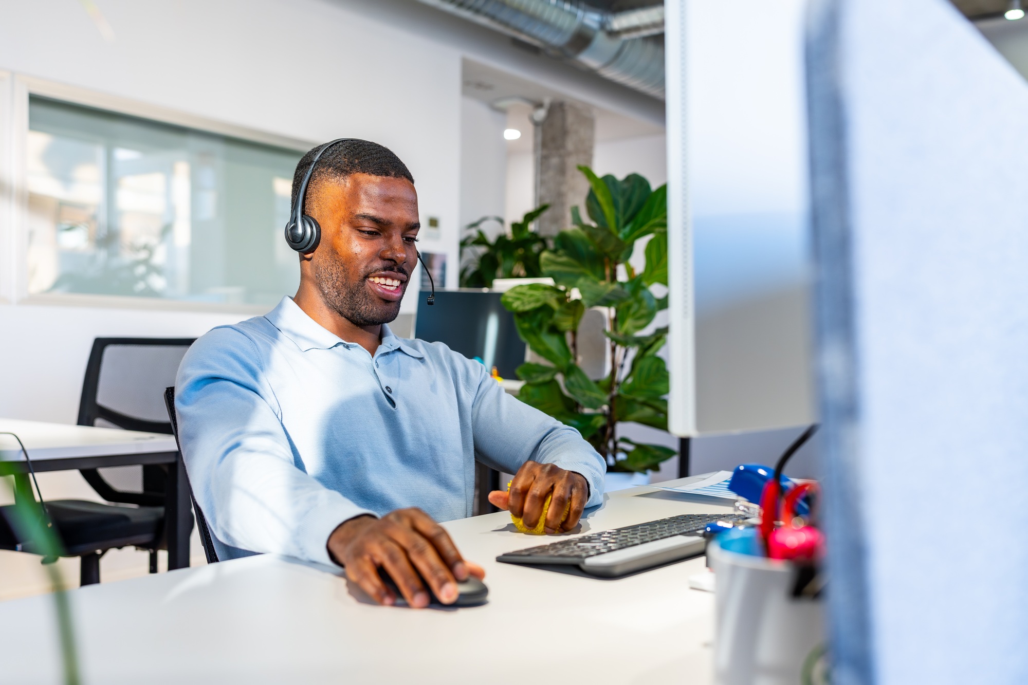 A professional using remote onboarding tools, wearing a headset and light blue shirt, works at a desk with a laptop in a modern office featuring plants and glass walls.