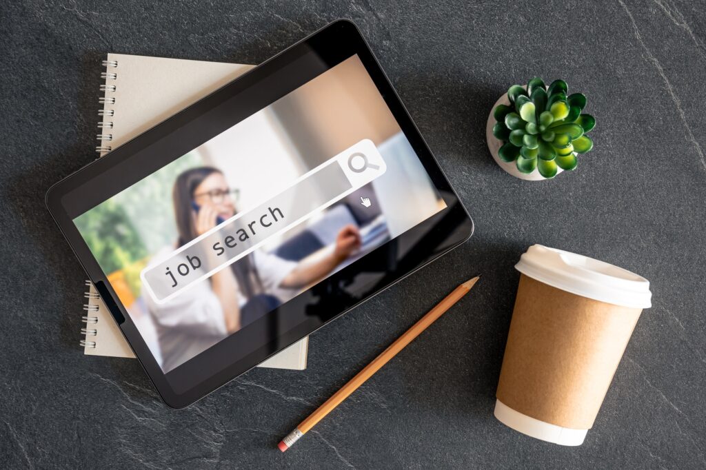 Tablet displaying a job search screen on a desk with a notepad, pencil, coffee cup, and plant—illustrating modern digital tools used in career placement services.