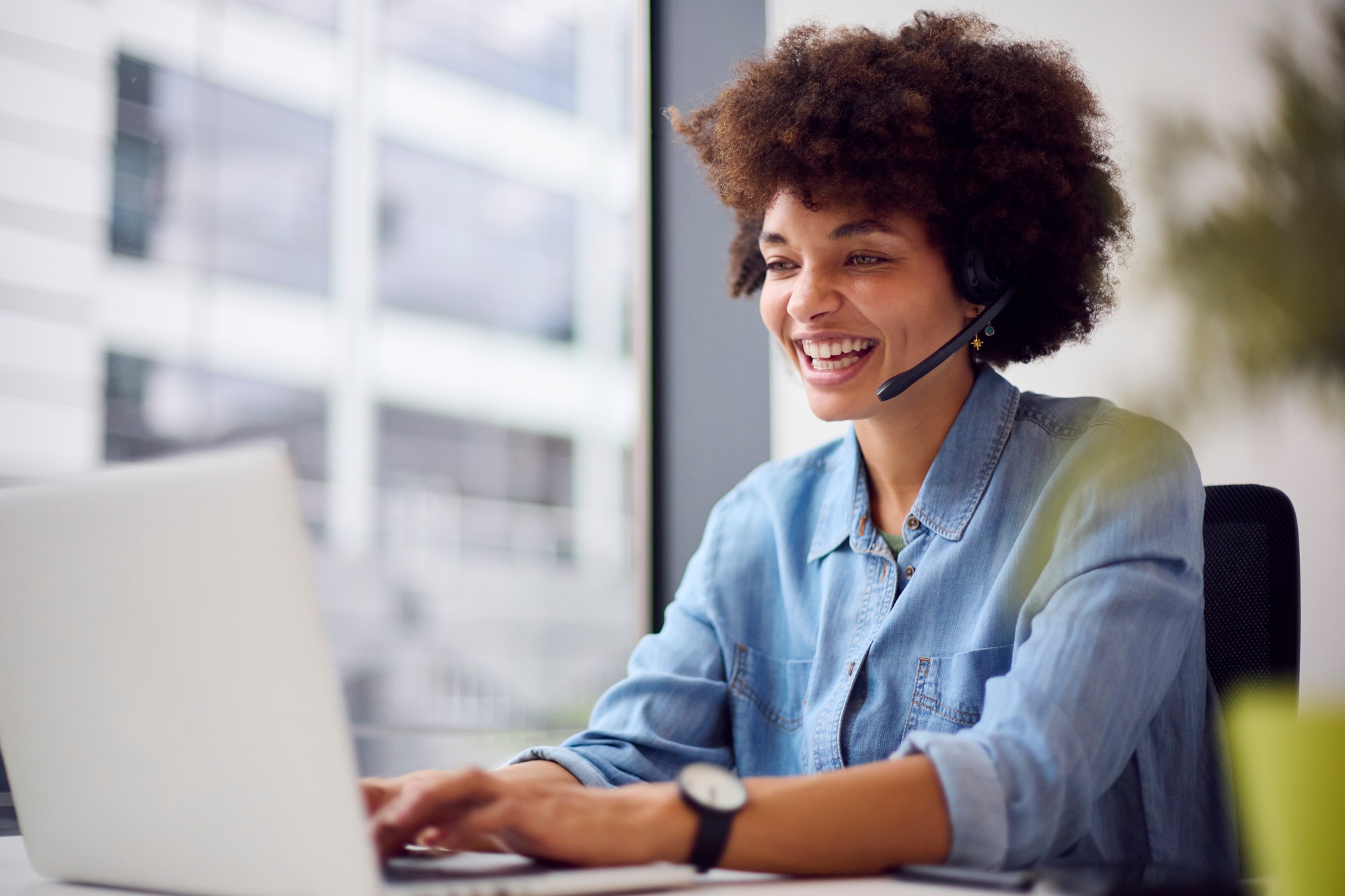 A professional providing virtual office support, wearing a denim shirt and headset, works at a desk with a laptop in a modern office with large windows and greenery in the background.